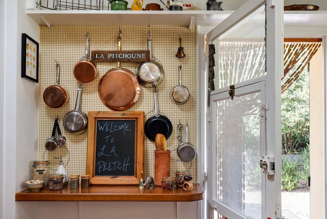 A kitchen interior with hanging copper pans, a chalkboard reading “Welcome to La Peetch,” and an open door to the outsid